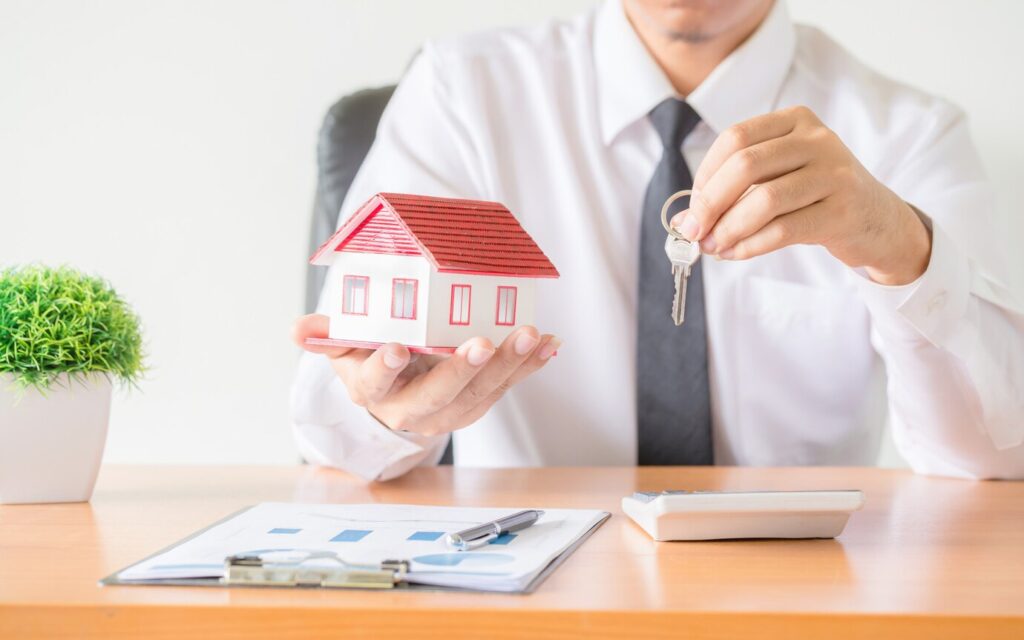 Man holding a model house, keys, and a clipboard of reports—an image symbolizing the management and oversight of Real Estate Investment Trusts (REITs).